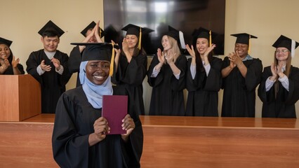 Happy African woman in hijab receiving diploma and classmates applauding her. 