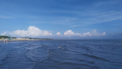 On the beach there are buildings, pine trees and a pier. The sandy beach has awnings, umbrellas and...