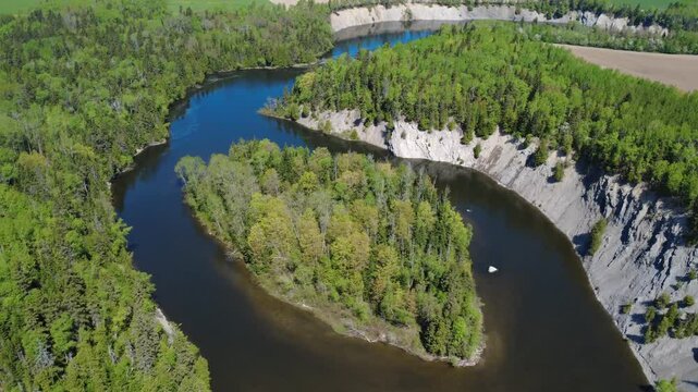Aerial drone view of an island in the center of a river near a steep cliff topped with forest, where a farm field is plowed by a tractor in the distance. Mitis River, Price, Quebec, Canada.