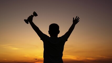 Child with champion cup raises hand rejoices in sports victory outdoors. Victory, child silhouette. Child athlete winner at sunset sky. Victory children dream. Best of best boy, achievement of success