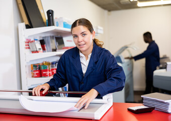 Positive female employee at printing house cuts paper with a paper cutter