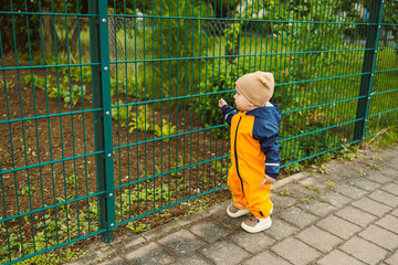 A curious toddler in a bright orange rain suit observes nature through a green fence, showcasing...