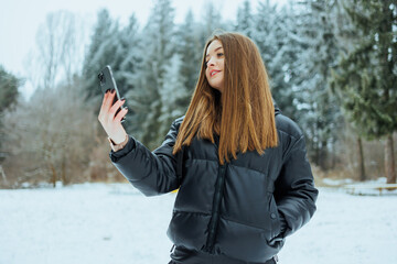 A young woman wearing a stylish winter coat is taking a cheerful selfie in a beautifully snowy...