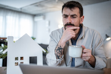 Thoughtful man drinking coffee and evaluating real estate investment on laptop