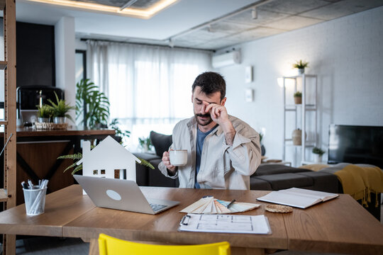 Tired architect rubbing eyes while drinking coffee at home office desk - Powered by Adobe
