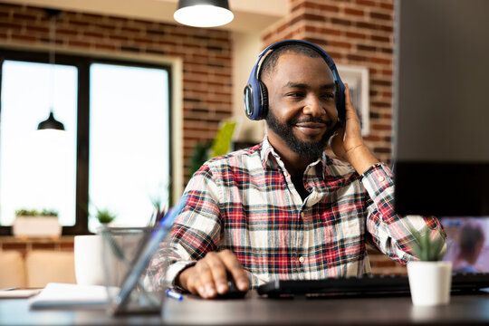 Male financial analyst seated at desk, listening to music and remotely reviewing company reports on computer. African american businessman works from home, using desktop pc and wireless headset.