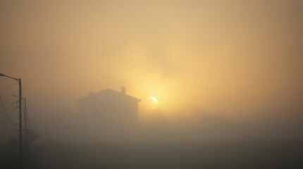 Obraz premium Misty Morning Silhouette: A serene early morning scene unveils a house shrouded in a thick fog, where the rising sun bathes the landscape in a soft, golden glow.