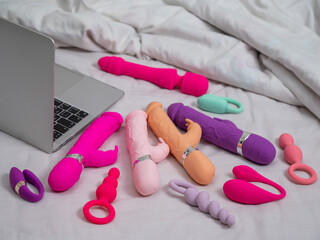 Collection of sex toys on a white sheet. Various vibrators and a laptop on the bed. 