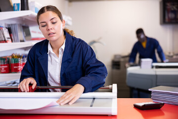 Focused young female specialist in uniform trimming the printed work on the cutter in the typography
