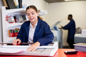 Cheerful young female specialist in uniform calculates the cost of notepads and sheets in the typography