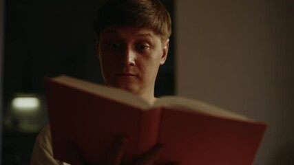 Close-up portrait of passionate young man reading paper book in dark bedroom, surrounded by warm light that enhances cozy and peaceful atmosphere of home. Concept of evening relaxing alone.