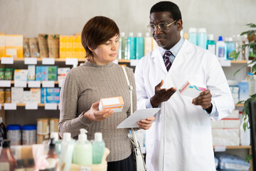 Adult woman buyer choosing box of pills with help of adult man pharmacist in pharmacy