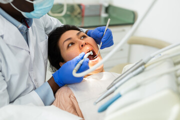 Asian woman sitting on dental chair with opened mouth and waiting for medical procedures ending in...