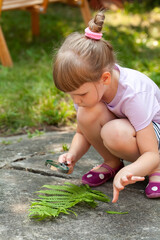 Young girl child crouching outdoors studying fern leaves using a magnifying glass exploring nature and learning about ecology and environment in summer backyard nature observation curiosity concept