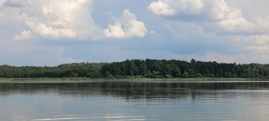 Wide, calm lake reflecting lush green forest and a cloudy sky on a summer afternoon, creating a tranquil and serene natural landscape filled with beauty and harmony
