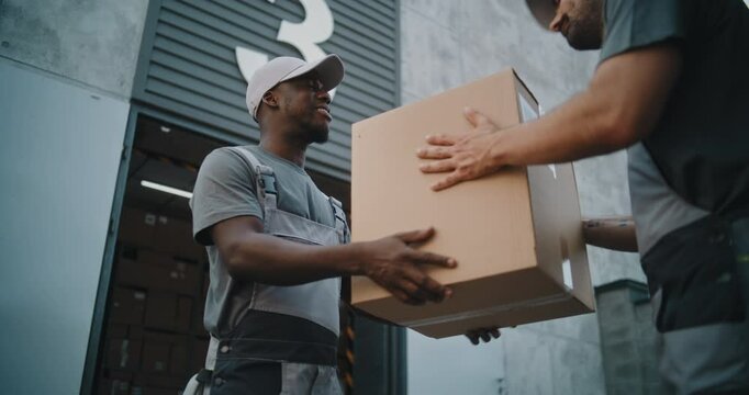 Outside of Logistics Distribution Warehouse: African American Worker Giving Cardboard Box with Online Orders, E-Commerce Goods to Colleague for Delivery. Working in Delivery Service, Storage Facility.