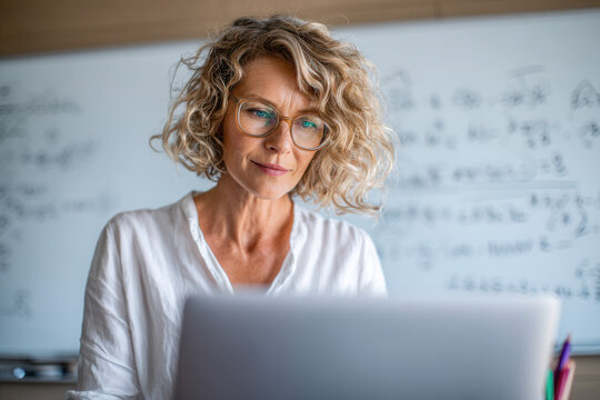 Middle-aged Caucasian woman with curly blonde hair wearing glasses, working on a laptop in a classroom with math equations on the board.