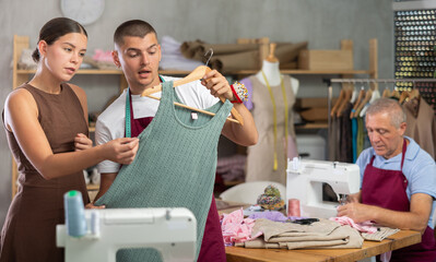 Tailor shows a woman a sewn dress against the background of an elderly employee of the atelier....