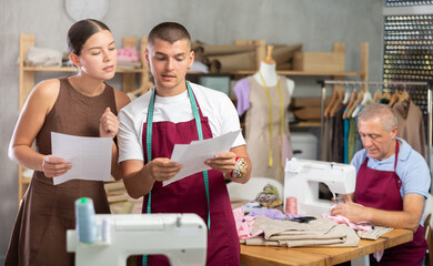 Guy tailor shows paper sketch of dress to girl client, demonstrate financial documents and explains cost of work, senior man dressmaker work with sewing machine in background.