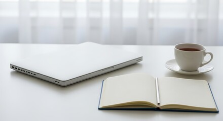 Fototapeta premium A minimalist workspace featuring a laptop notebook and a cup of tea on a white table near a window with soft natural lighting perfect for work or relaxation.