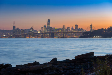 Fototapeta premium San Francisco, USA: The city skyline and Bay Bridge are illuminated at dusk. The lights of the buildings and bridge reflect on the water.