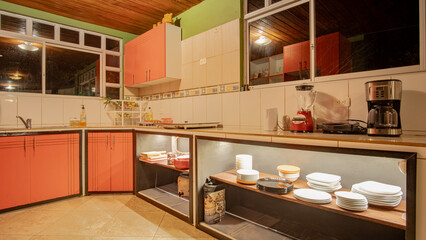 Warm rustic kitchen with red cabinets, open wood shelves displaying plates and bowls, cozy nighttime lighting, Ecuador.