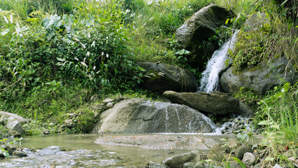 Small natural waterfall cascading over large rocks into a clear shallow stream surrounded by dense...