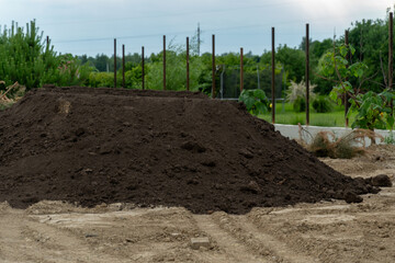 A sizable pile of dark soil sits on a farm landscape surrounded by patches of grass and emerging...
