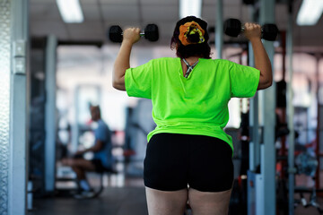 Beautiful overweight Latina woman lifting dumbbells in a gym, wearing a green 80s-style t-shirt,...