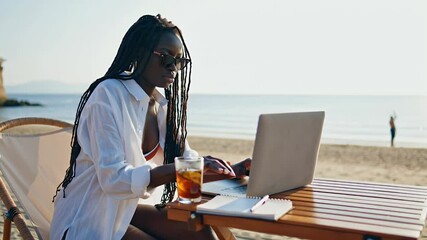 Young black woman working remotely on laptop computer at beach with iced tea during summer vacation trip - Powered by Adobe
