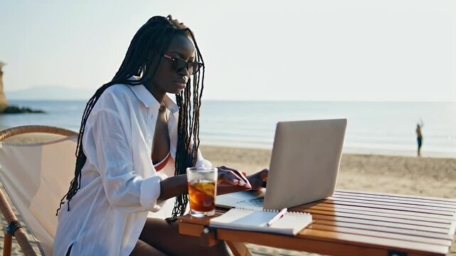 Young black woman working remotely on laptop computer at beach with iced tea during summer vacation trip - Powered by Adobe