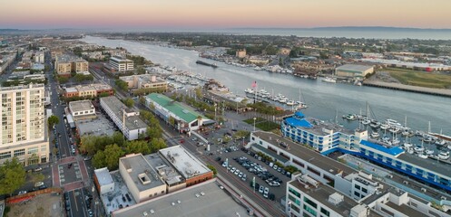Fototapeta premium Aerial view of ack London Square, Oakland, California, USA, showing the Oakland-Alameda Estuary, buildings, parking lots, and streets at dusk. The image captures the urban landscape.
