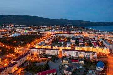 Beautiful night cityscape. Aerial view of a seaside town. Top view of illuminated streets and buildings. Mountains and sea in the distance. City of Magadan, Magadan region, Far East of Russia.