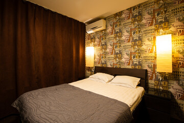 Apartment interior. Bed in the bedroom. Pillows and bedspread on the made bed. Wallpaper with images of European landmarks. Air conditioning and wall lamps on the wall. Windows covered with curtains.