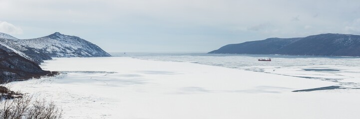 Cargo ship among ice floes in frozen sea bay. Mountains on coast. Panorama of Nagaev Bay, Sea of ​​Okhotsk, Magadan Region, Russian Far East.