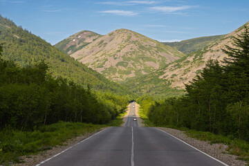 A beautiful road stretching into the distance between green hills and mountains. The asphalt road is surrounded by dense forest. Cars are driving along the road in the distance. Magadan Region, Russia