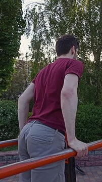 Young man doing dips on parallel bars, viewed from behind. Wearing a burgundy T-shirt and grey pants. Summer outdoor fitness workout.