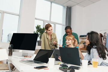 young adult coworkers collaborating in modern workplace with laptops and positive socializing,...
