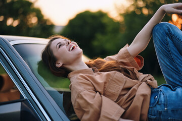 Young woman laughing joyfully on a car hood during sunset, wearing a casual brown shirt and blue...