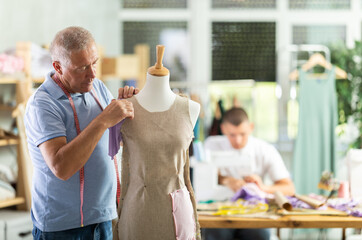 Elderly tailor checks a sample of a sewn dress on a mannequin against the background of a young...
