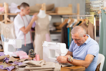 Mature tailor and a young assistant work in a sewing workshop, they sew clothes. Tailor's...