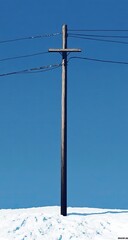 A weathered wooden utility pole, topped with a cross-shaped support, stands tall against a clear, bright blue sky.  It's positioned on a snow-covered hill