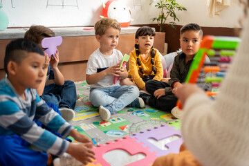 Group of preschool children sitting on the classroom floor while the teacher uses an abacus to...