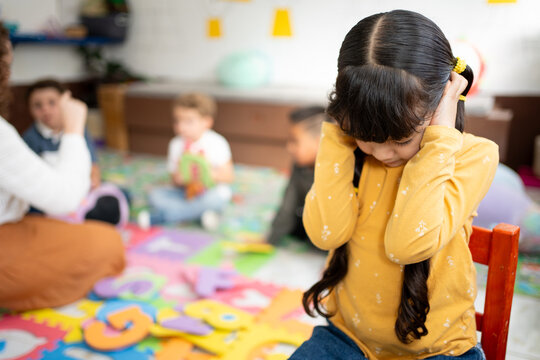 Preschool girl covers her ears and closes her eyes while other kindergarten children play loudly behind her. Concept of noise sensitivity, sensory overload, and emotional response in early childhood