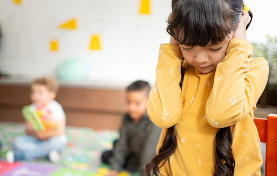 Preschool girl covers her ears and closes her eyes while other kindergarten children play loudly behind her. Concept of noise sensitivity, sensory overload, and emotional response in early childhood