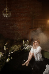 Young Caucasian woman, mid-20s, light brown hair, in white dress, front view, sits in shallow artificial pond with fog and flowers, brick wall behind. Concepts: fantasy, nature, calm, dreamlike.