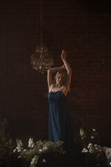 Young Caucasian woman in blue dress posing with raised arms, looking away. Front view. Vintage room with brick wall, crystal chandelier, flowers. Concepts: elegance, beauty, nostalgia, romance.