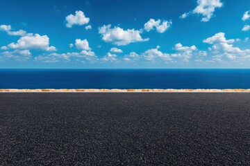 Obraz premium Wide asphalt road stretching towards the horizon, bordering a vast blue ocean under a sunny sky dotted with white clouds, creating a scenic coastal drive