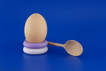 Egg balanced on stacked soap rings alongside a wooden spoon on a blue backdrop.