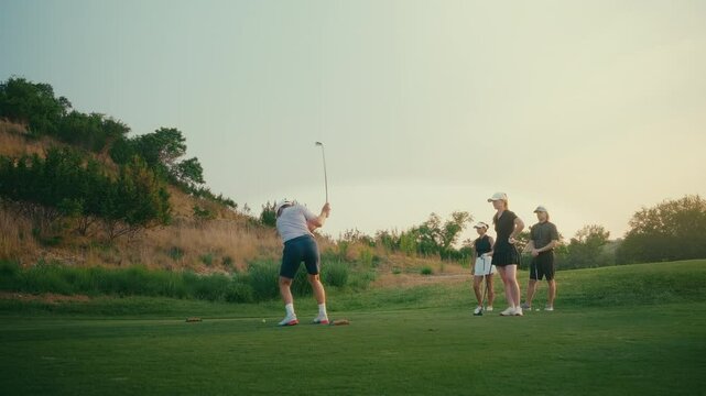 Golf foursome gathers on the tee box in golden hour light as a male golfer hits an iron shot. After the swing, the group celebrates with a cheerful high five in a relaxed, scenic moment.